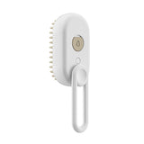 A white electric hairbrush with a hanging loop handle, featuring a circular gold-toned power button with a water droplet icon on the front.